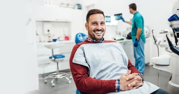 Adult Male in red sweater sitting in a dental chair with a paper draped over his chest and is smiling at the camera